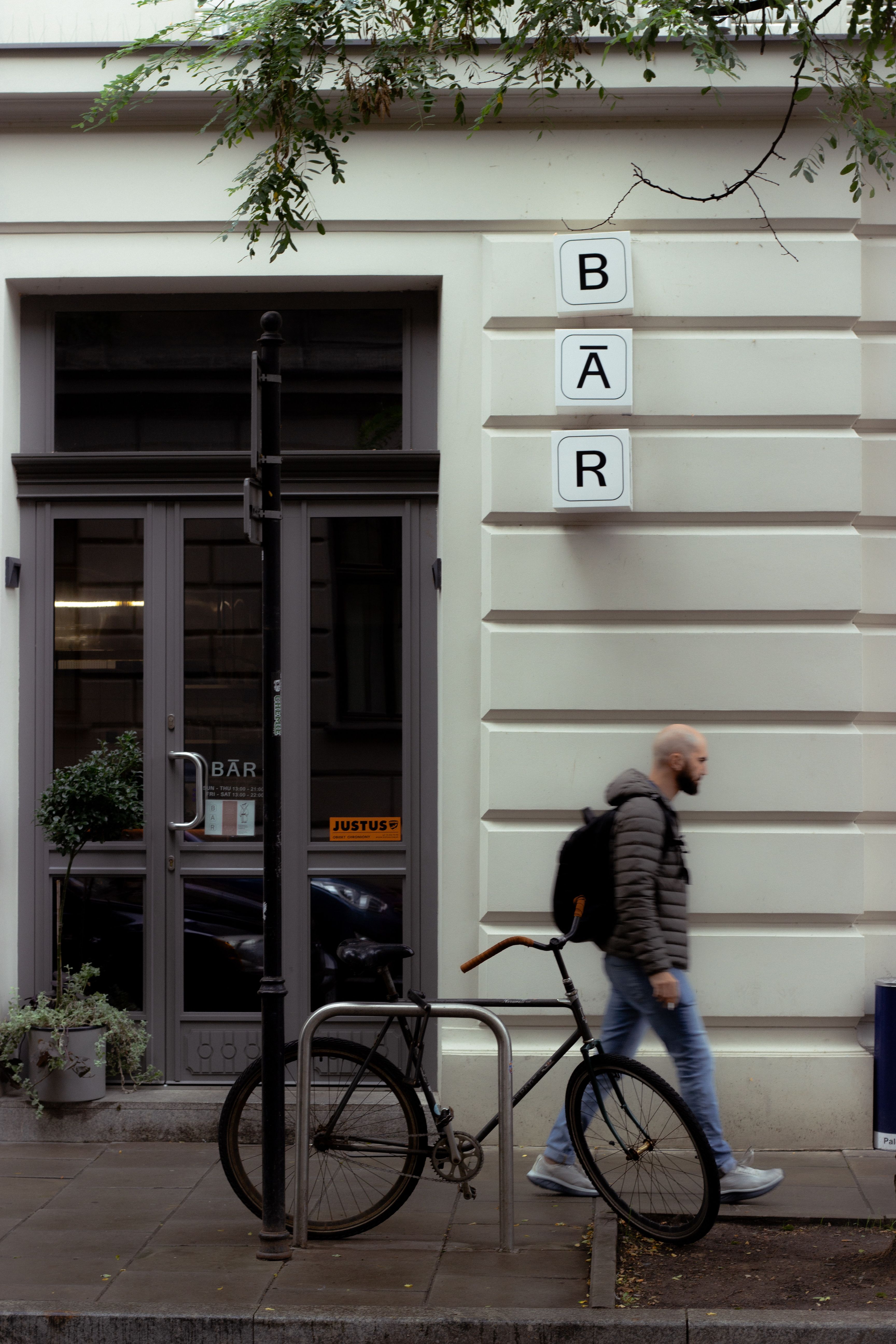 A person walks by a bar entrance in a city street with a bicycle parked nearby.