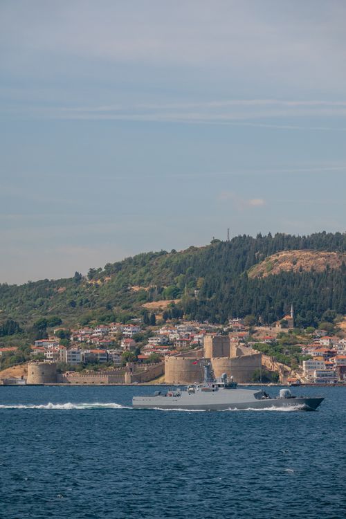 Free A naval ship navigates the waters near a historic stone fortress with a lush, hilly backdrop. Stock Photo