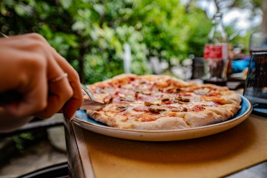 A tempting Italian pizza being sliced, served outdoors with vibrant greenery.