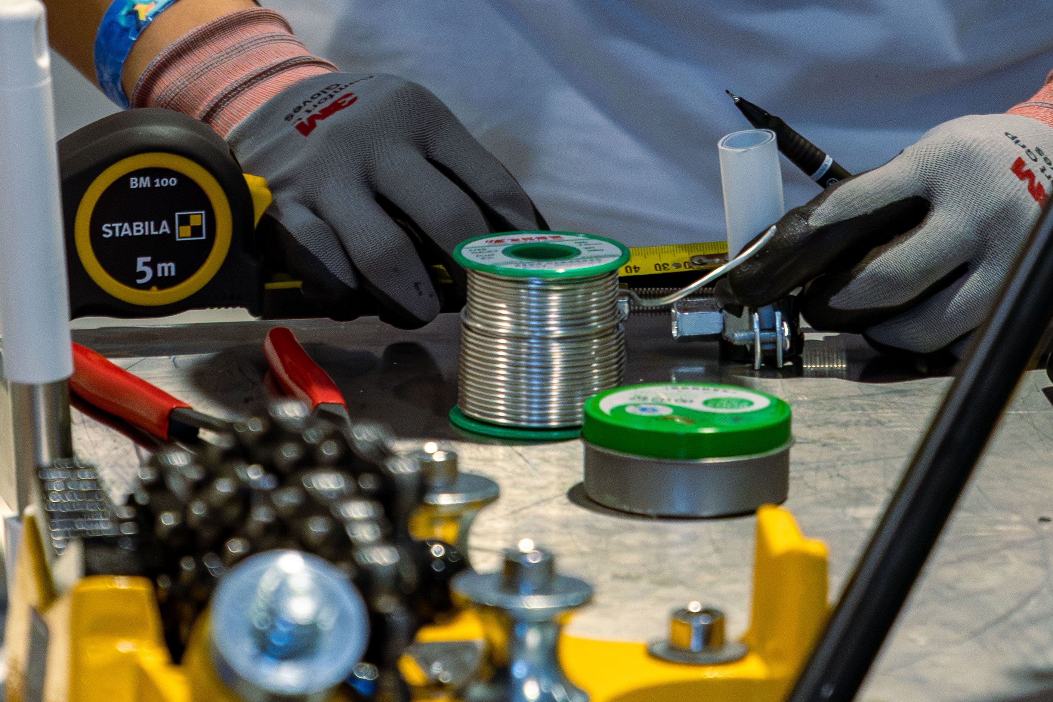 Detail view of workshop tools, including tape measure and soldering materials, handled with precision gloves.