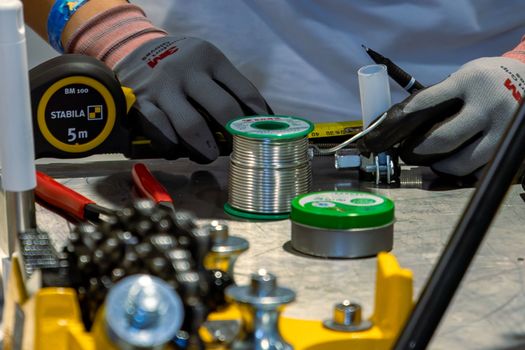 Detail view of workshop tools, including tape measure and soldering materials, handled with precision gloves.