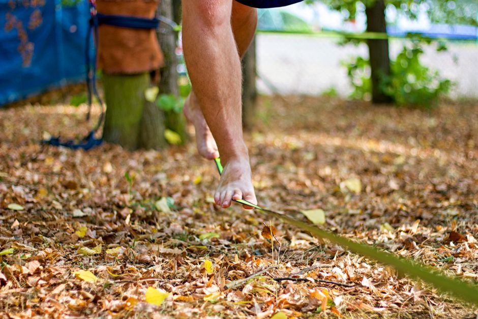 A person balances barefoot on a slackline surrounded by autumn leaves in a park setting.