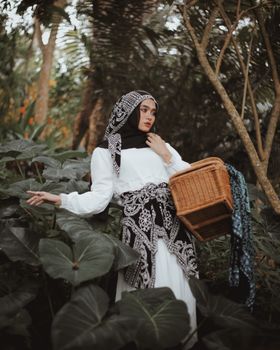 一位头戴头巾、身着时尚服装的年轻女子，在户外将一个篮子摆放在郁郁葱葱的绿色植物环绕之中
