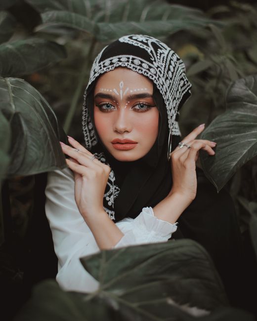 A sophisticated portrait of a woman in a hijab with intricate makeup, surrounded by lush greenery.