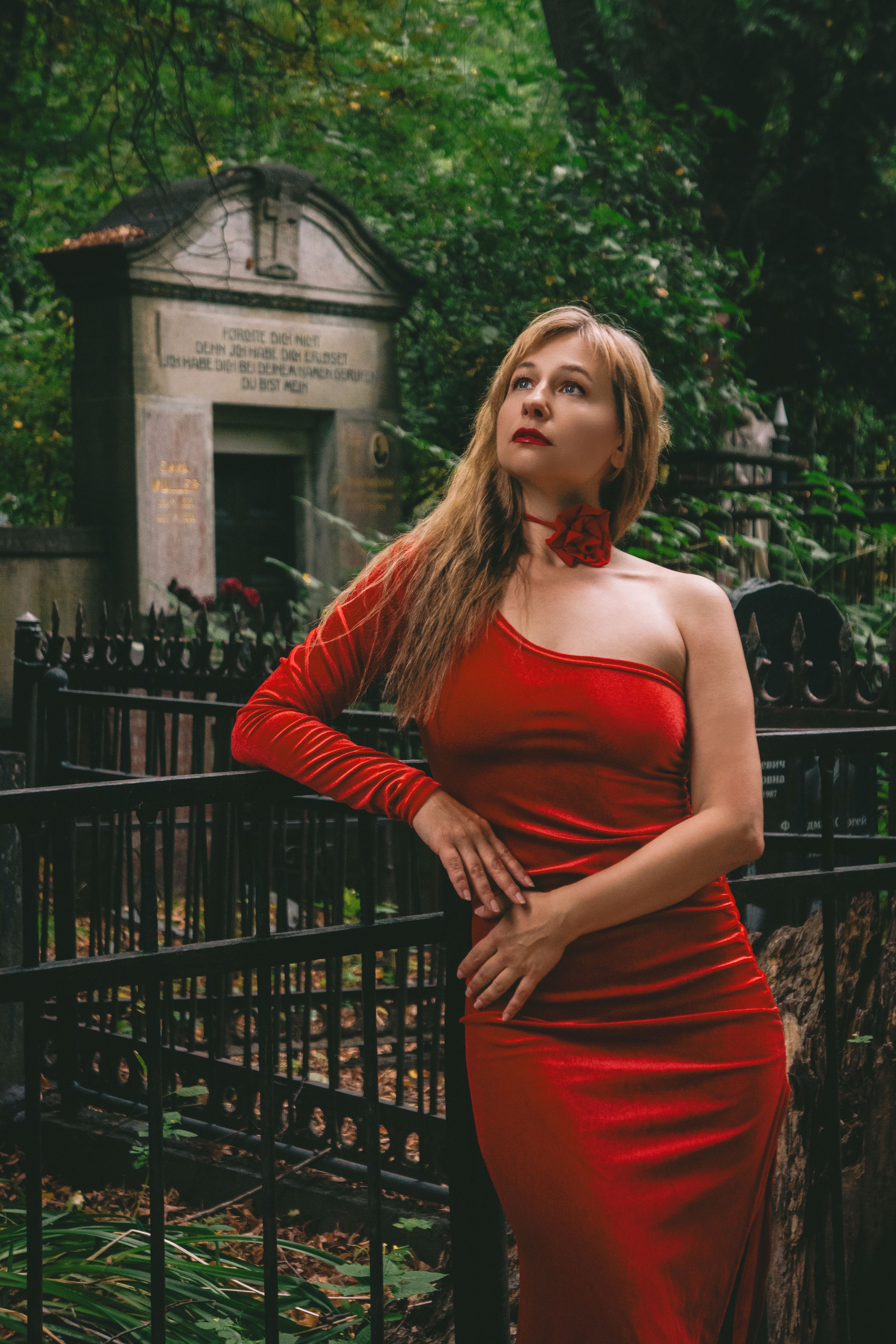 Elegant woman in a red dress stands by a gothic tombstone in a historical cemetery.