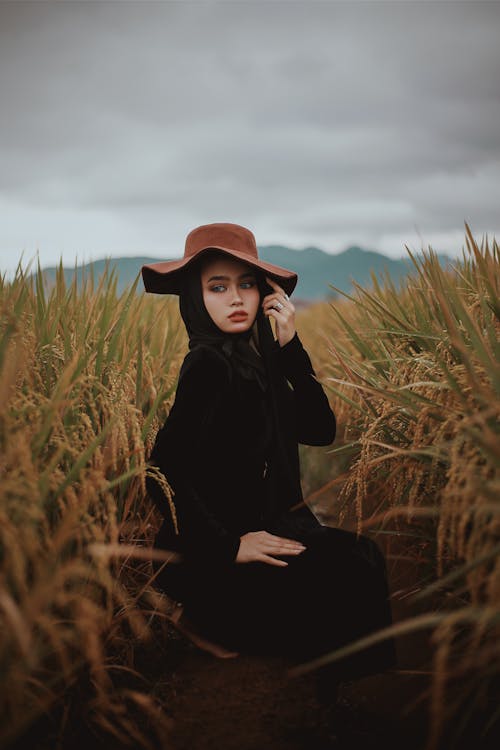 Woman in Black Long Sleeve Shirt and Brown Hat Sitting on Brown Grass Field