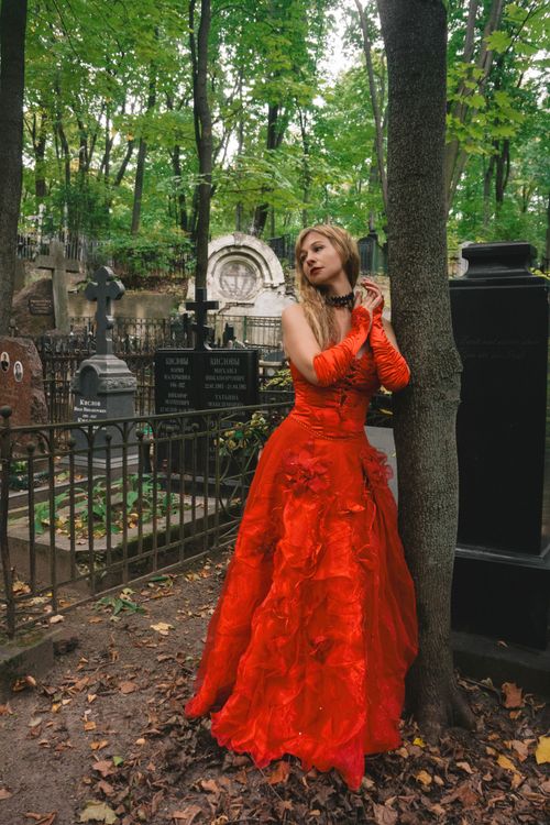 Free A woman in a vibrant red gown poses in a historic cemetery, surrounded by lush green trees. Stock Photo