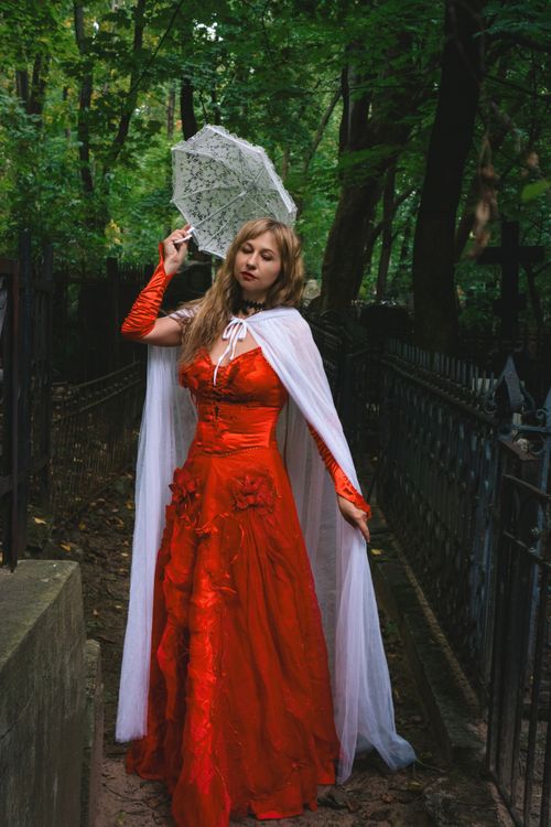 Free A woman in a striking red dress poses outdoors holding a lace umbrella amidst a forest setting. Stock Photo