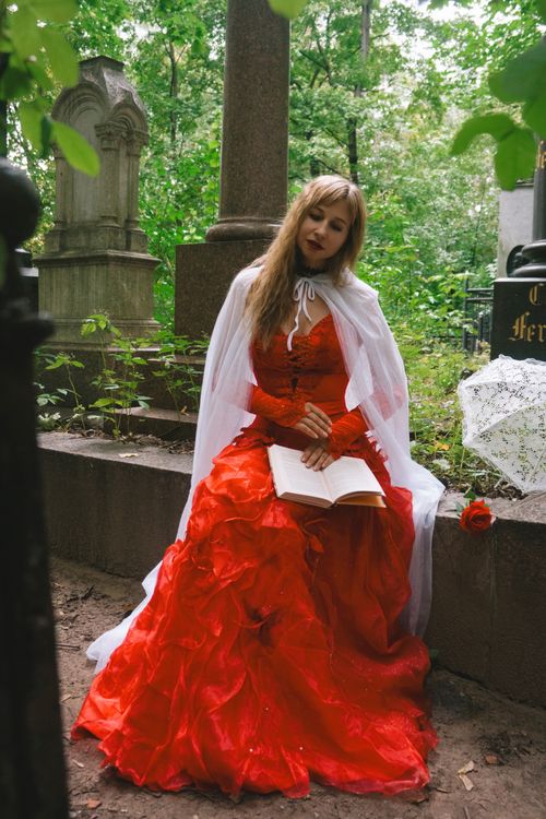 Free Woman in a red dress reads solemnly in a gothic cemetery setting, surrounded by greenery. Stock Photo