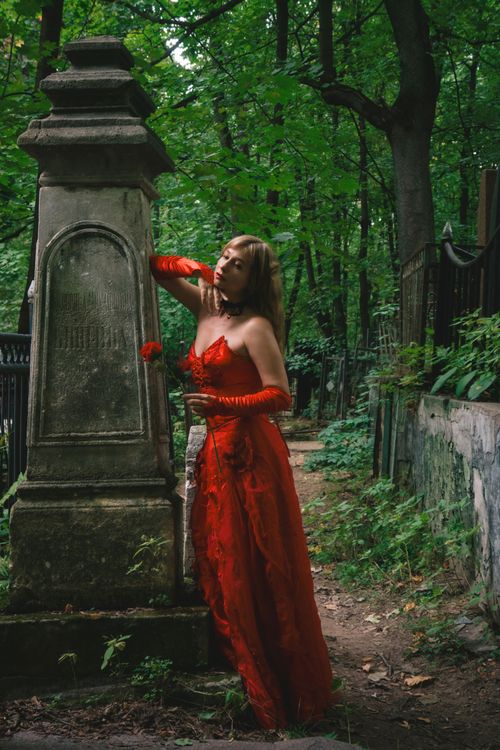 Free A woman in a striking red dress poses dramatically beside an old tombstone in a lush, green cemetery setting. Stock Photo