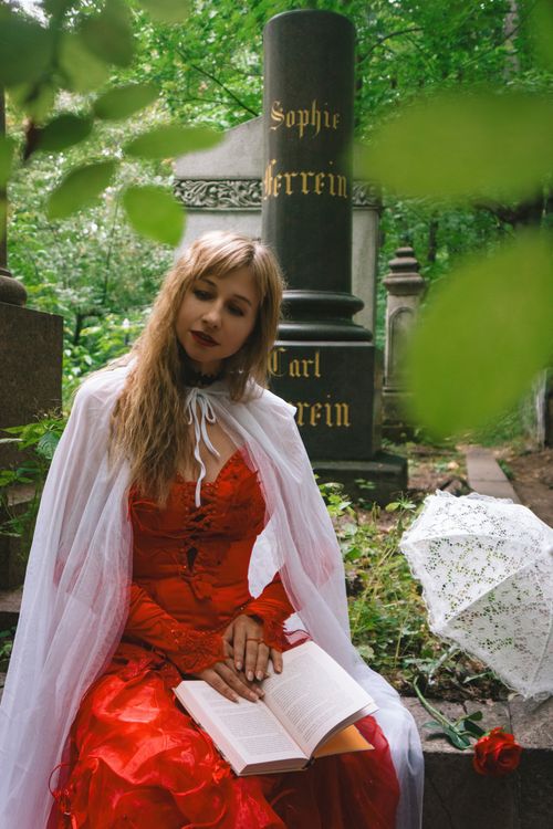 Free Woman dressed in red, reading in a serene cemetery setting, evoking mystery and solitude. Stock Photo
