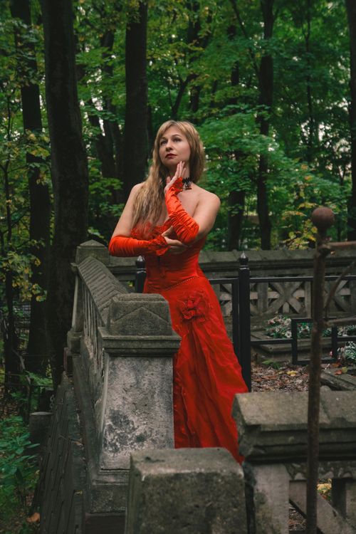 Free Elegant woman in a red gown stands by a stone fence in a lush green forest setting. Stock Photo