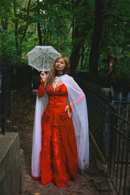 Free A woman wearing a red dress and white cape holds a lace umbrella while standing in a lush green setting. Stock Photo