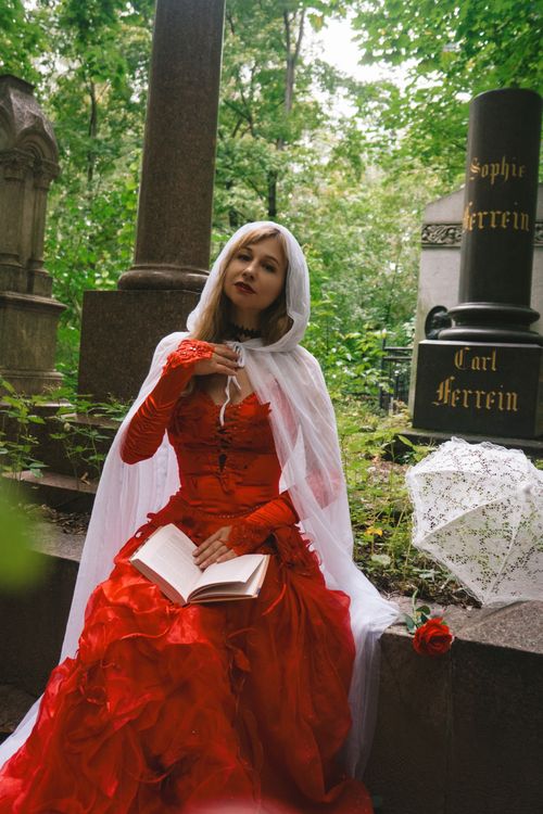 Free A woman in a vibrant red dress reads an open book sitting in a lush, green cemetery next to a gravestone. Stock Photo