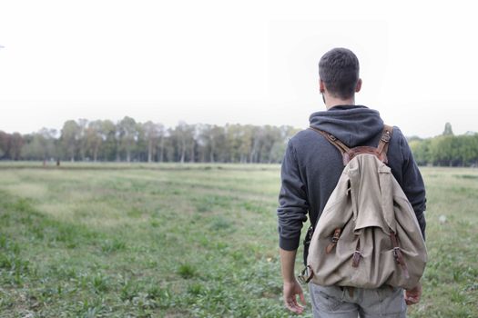 A man with a backpack walks through a vast open field surrounded by greenery and trees.