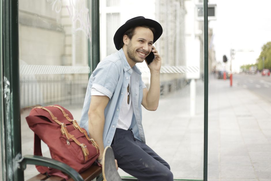 Casual young man sitting at a bus stop, using phone, and smiling in an urban environment.