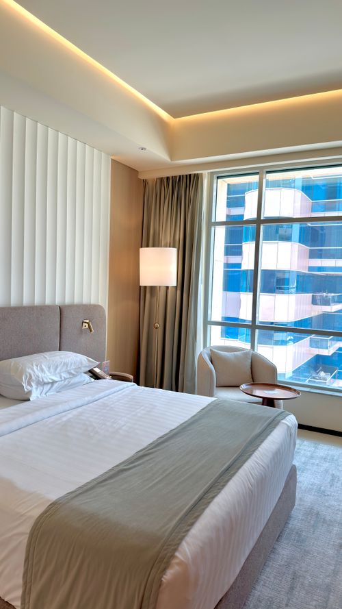 Free Elegant hotel room featuring a cozy bed, modern decor, and a city view through a large window. Stock Photo