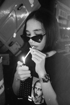 Black and white of young trendy ethnic female millennial in stylish outfit and sunglasses lighting cigarette