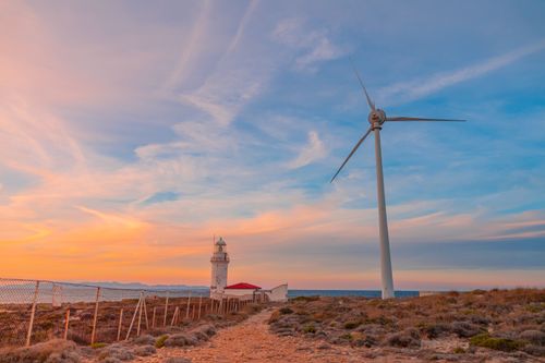 Free A serene image of a wind turbine and lighthouse by the coast during sunset, highlighting renewable energy. Stock Photo