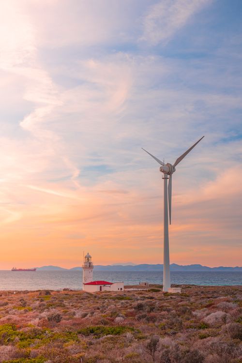 Free Wind turbine and lighthouse by the sea under a colorful sunset sky, symbolizing renewable energy. Stock Photo