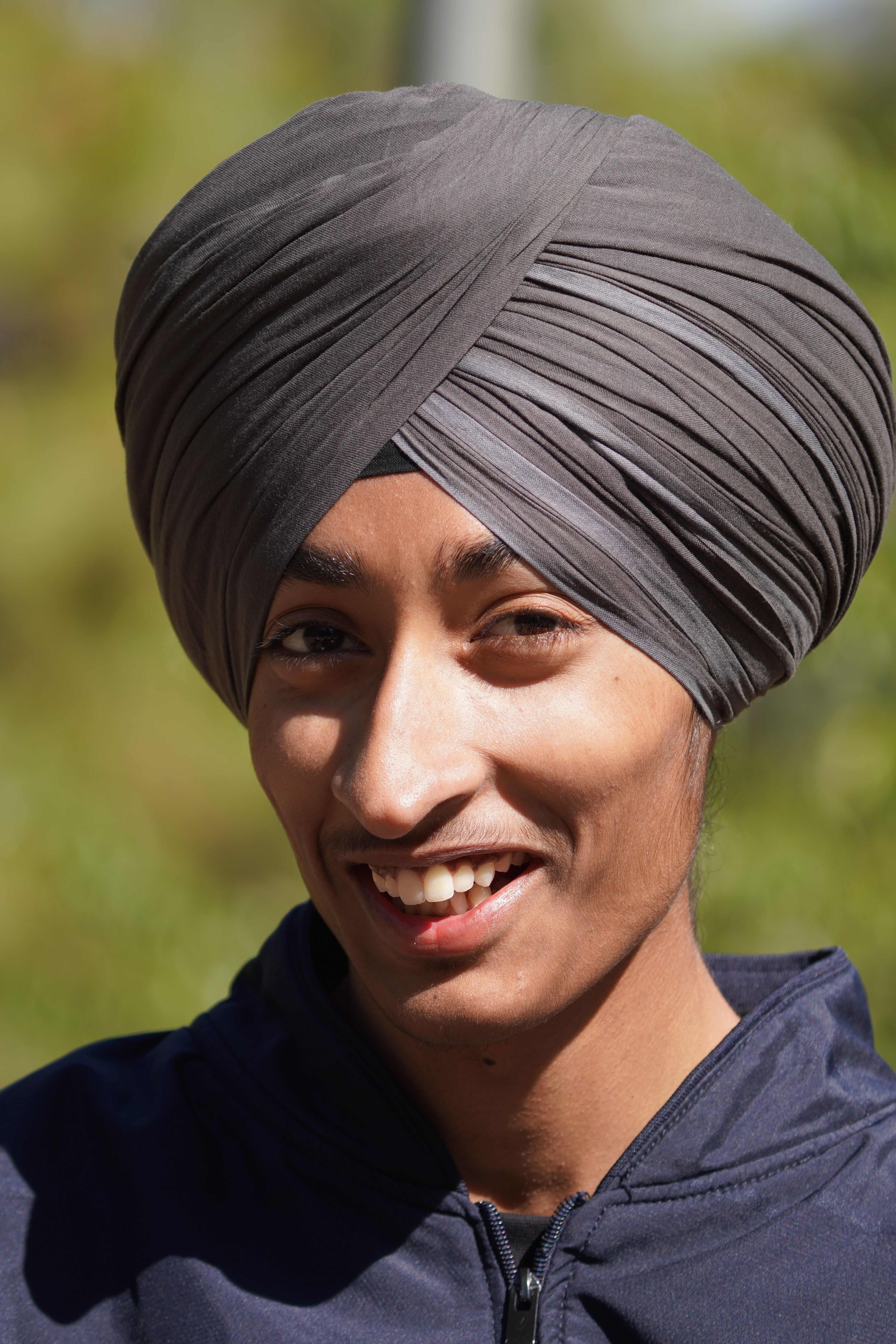 A cheerful young man wearing a turban smiles warmly outdoors, surrounded by greenery.