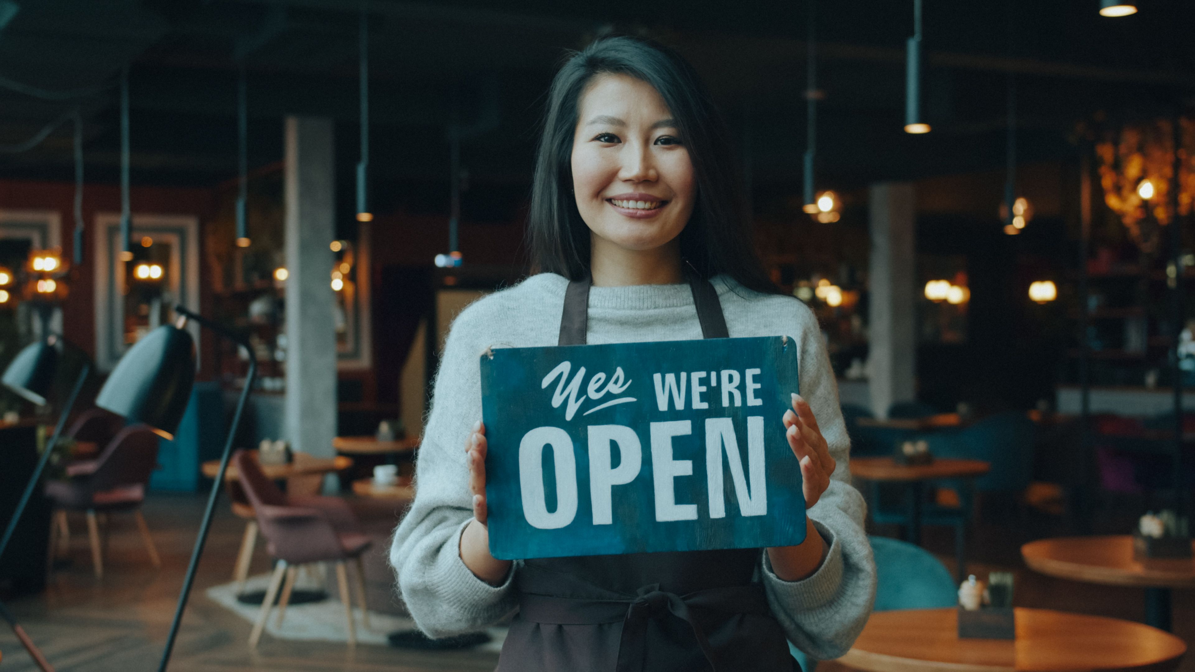 A smiling cafe owner holding an open sign inside a small restaurant