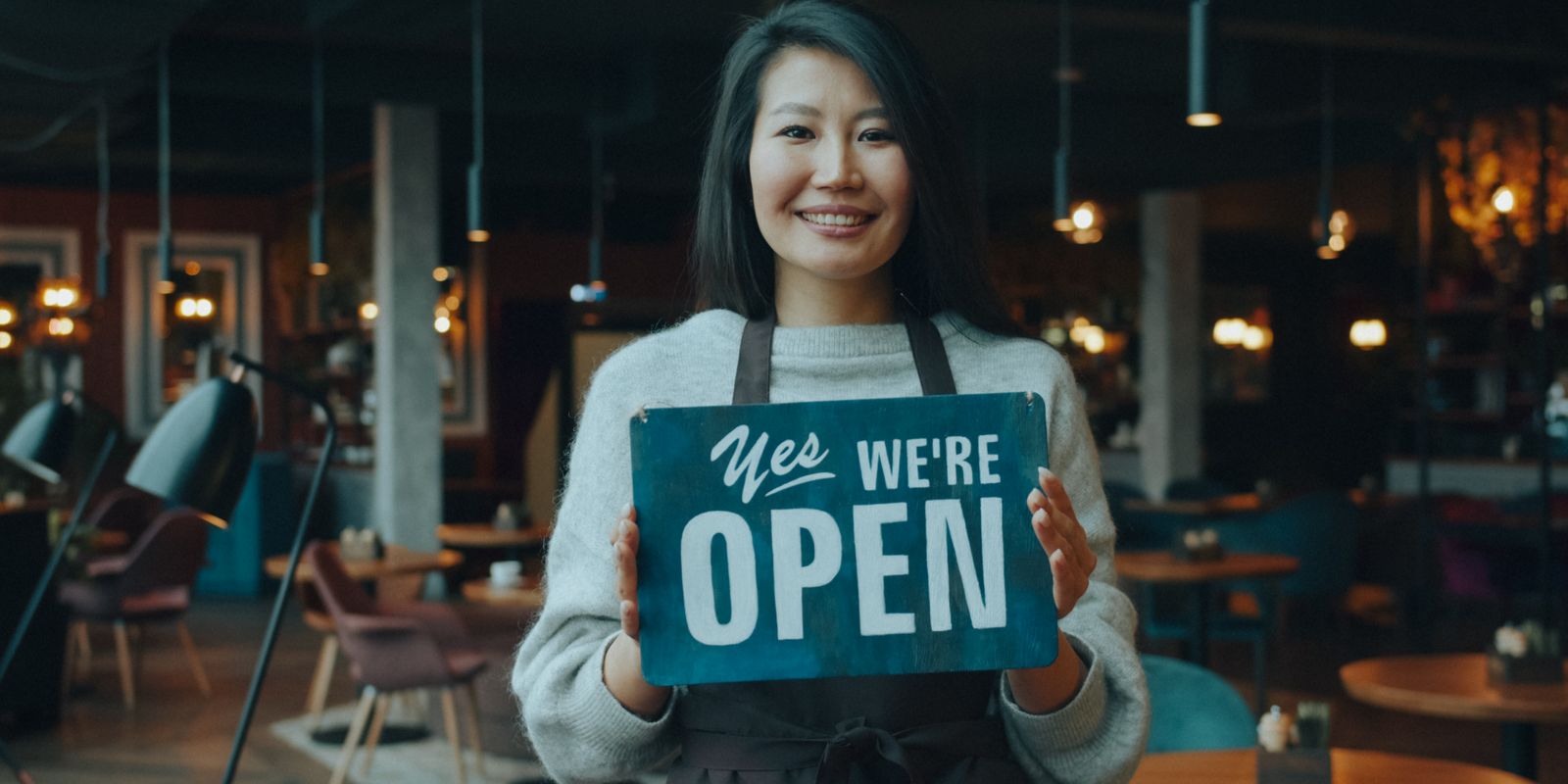A smiling cafe owner holding an open sign inside a small restaurant