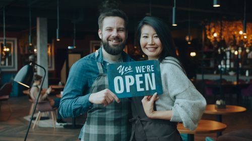 Free Smiling cafe owners holding an open sign inside their cozy cafe, welcoming customers warmly. Stock Photo