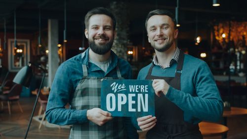 Free Two cafe owners smiling and holding a 'Yes We're Open' sign inside their cozy cafe. Stock Photo