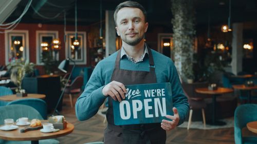 Free Cafe owner standing inside, holding a 'We're Open' sign, welcoming customers. Stock Photo