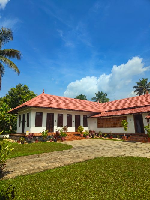 Free Charming traditional Kerala house with a red tiled roof and wooden pillars, surrounded by lush greenery. Stock Photo