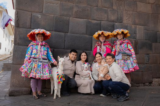 A family poses with Andean women in traditional colorful clothing, featuring alpacas, in a historic stone wall setting.