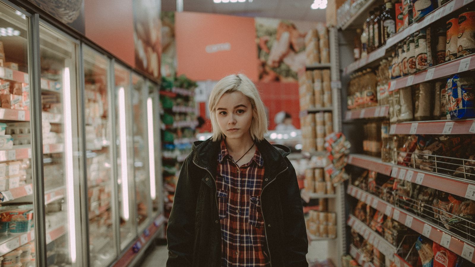 A woman with white hair stands between shelves in a supermarket aisle, showcasing a variety of products.