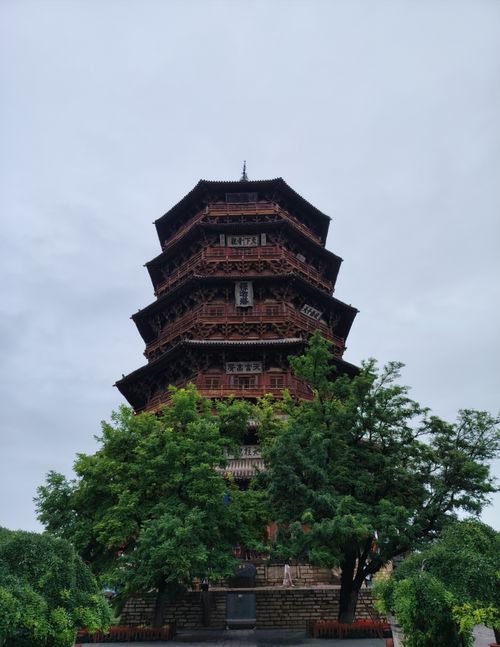 Free Front view of a historical wooden pagoda nestled among lush greenery under an overcast sky. Stock Photo