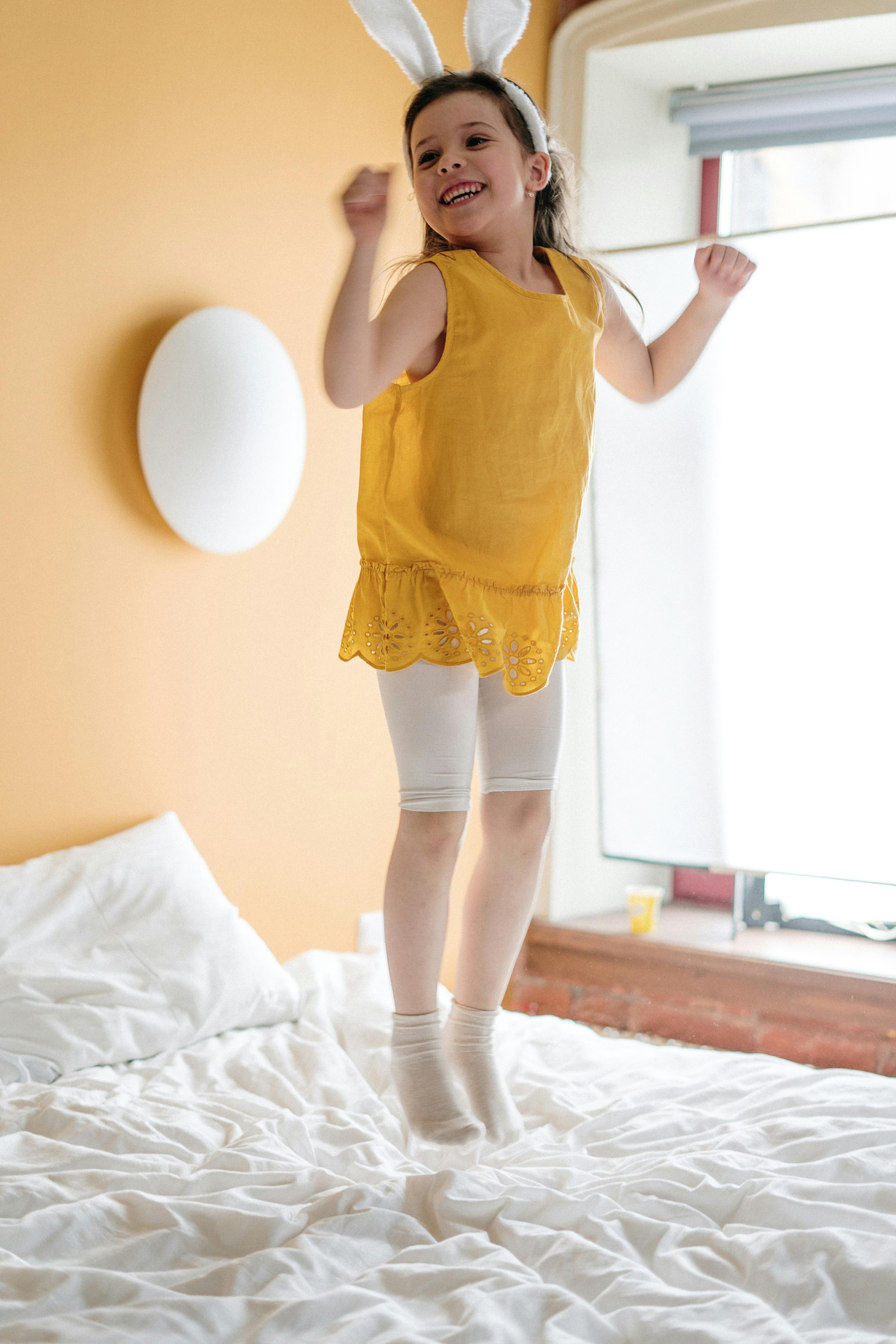 Young girl with bunny ears jumps on bed in a sunlit room, enjoying playful moments.