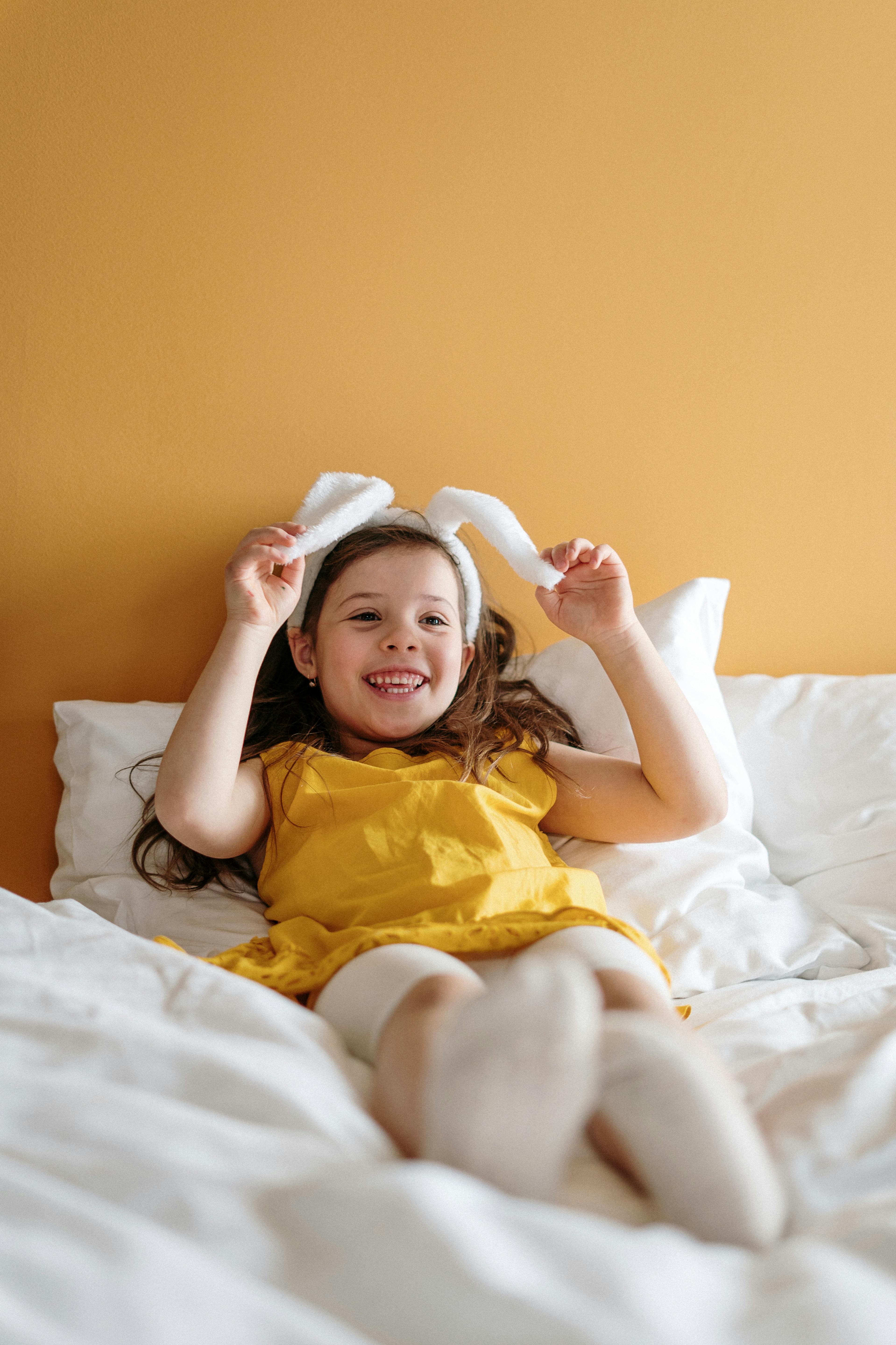 Smiling girl with bunny ears relaxing on a bed against a yellow background.