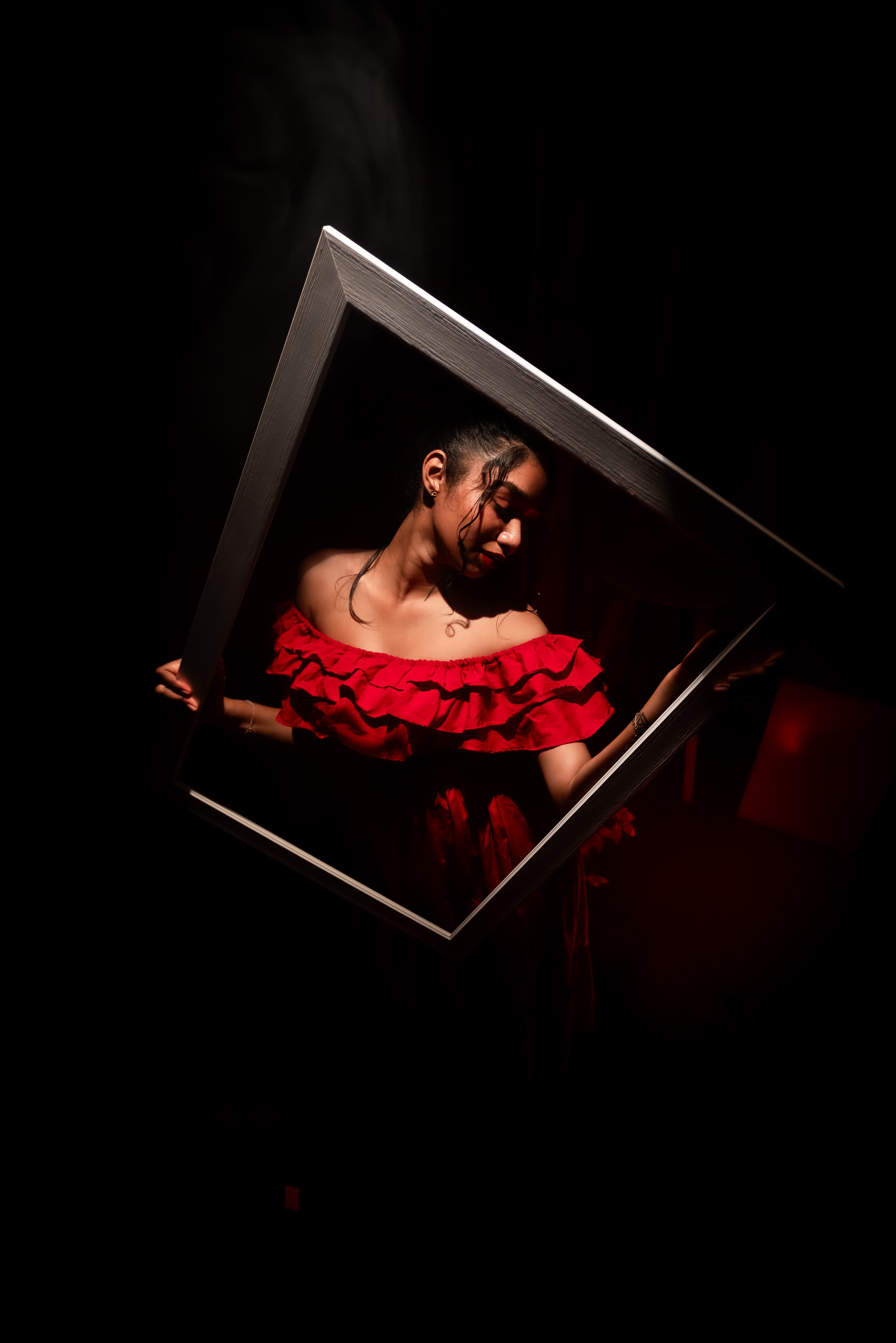 Portrait of a woman in red dress holding a frame, spotlighted in a dark setting.