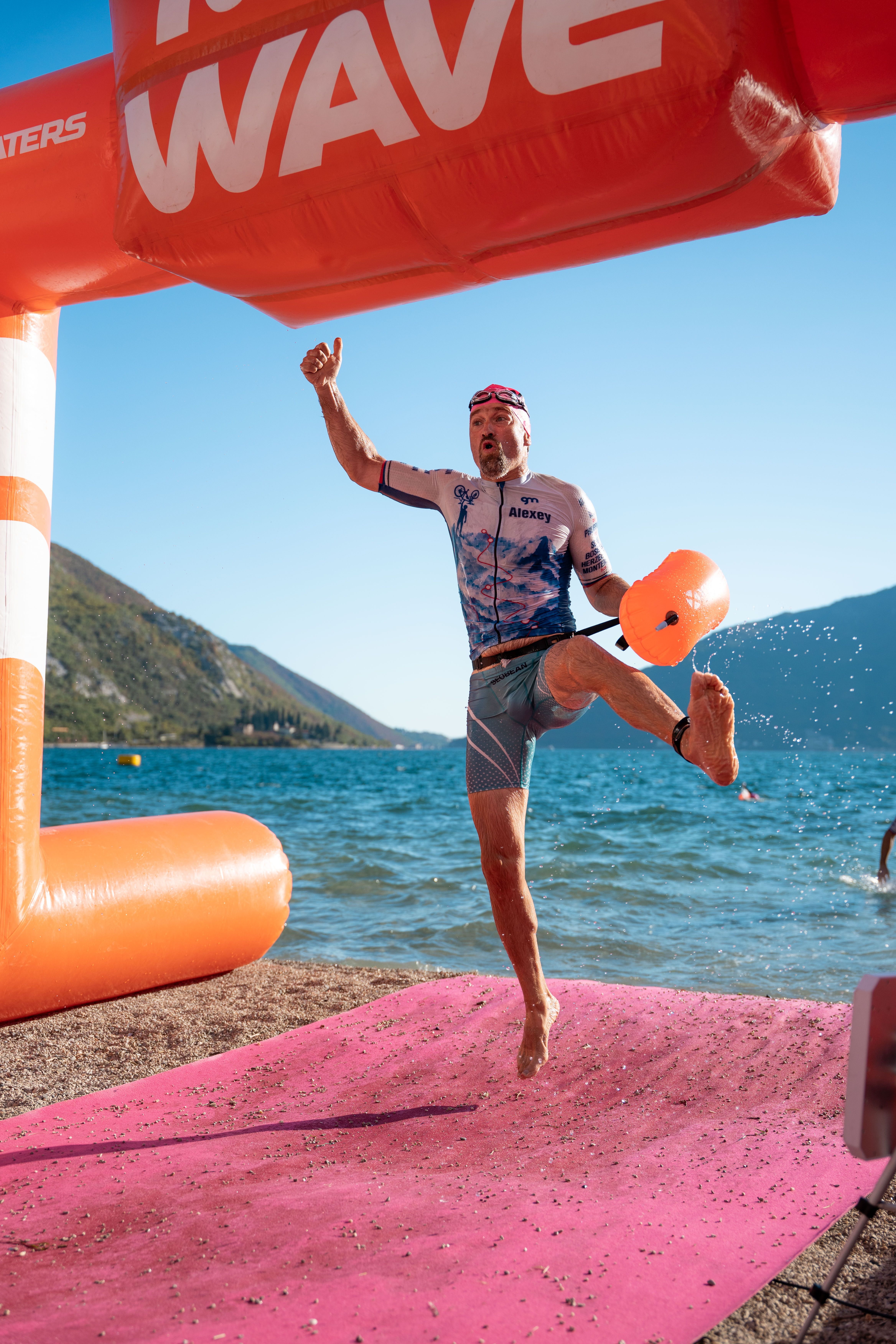 Triathlete leaps triumphantly across the finish line of a sunny beachside race under clear blue skies.