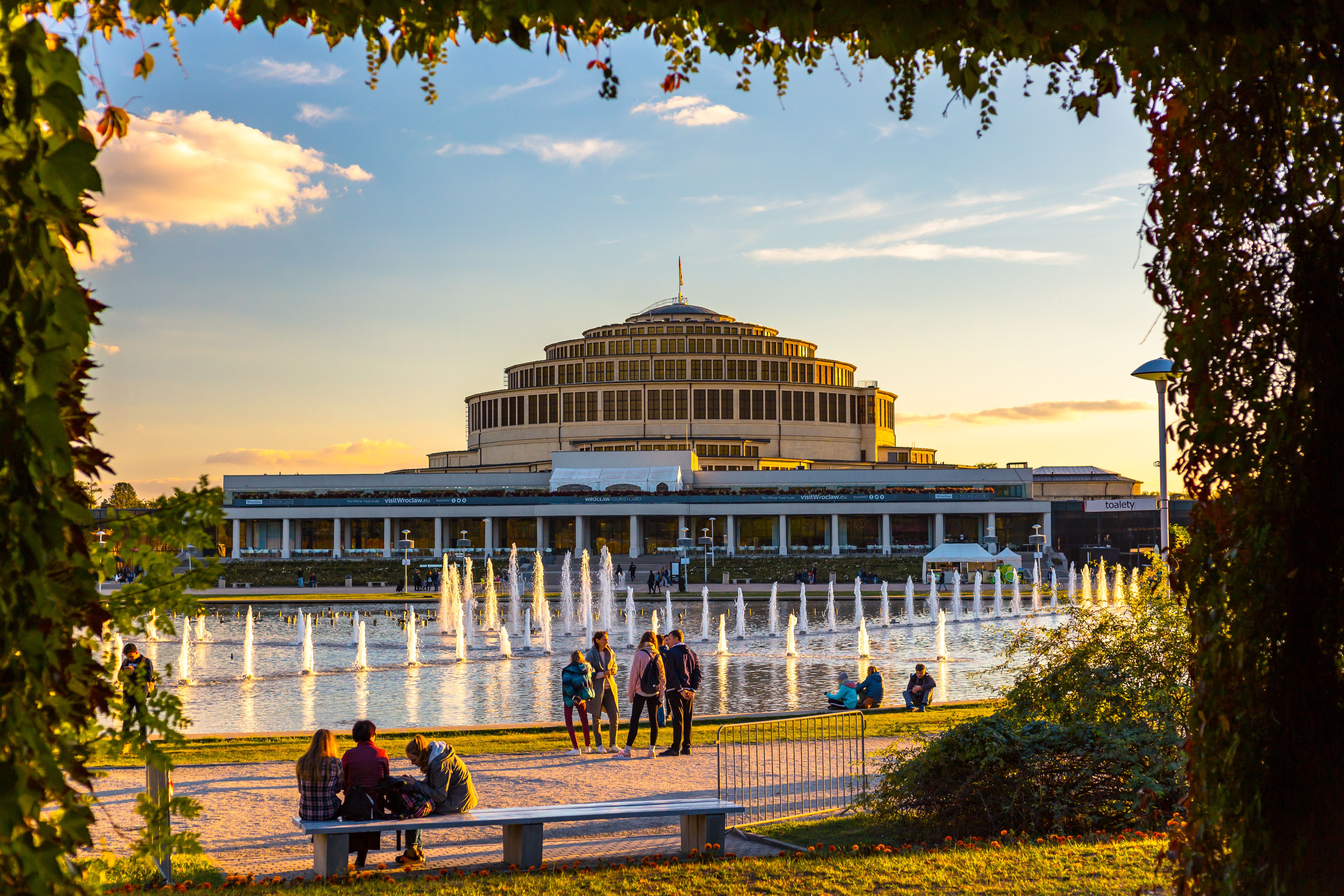 A scenic view of Wrocław's Centennial Hall surrounded by fountains and people enjoying sunset.