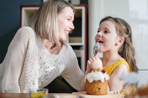 Free Woman in White Knit Sweater Smiling while Little Girl Licking Icing on Her Spoon Stock Photo