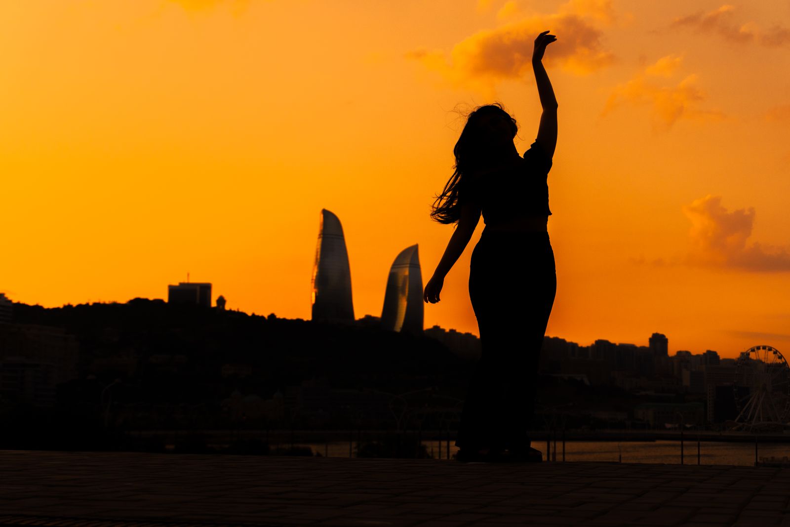 Silhouette of a woman with one arm raised against an orange sunset over a city skyline