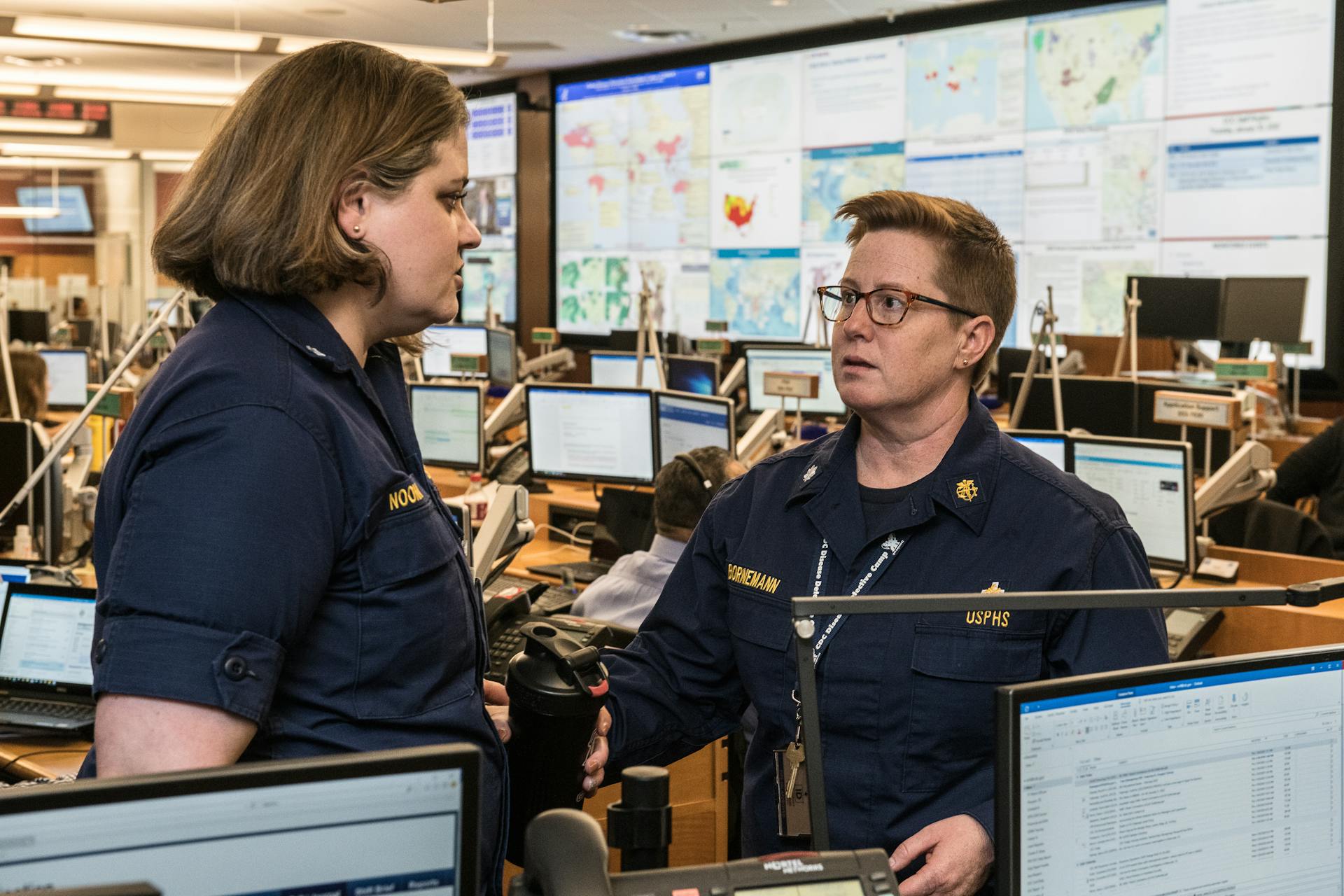 Adult and mature serious female colleagues in uniforms talking and looking at each other while standing near tables with computers behind stand with maps in modern office