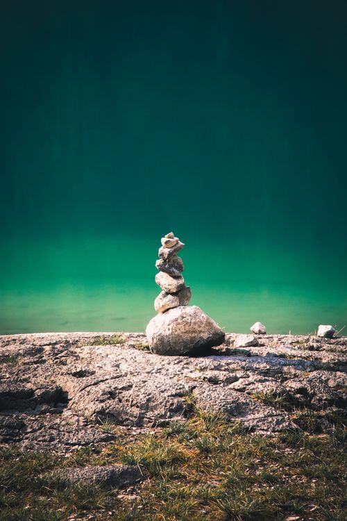 Free A tranquil stone cairn stacks elegantly near a serene, turquoise lake under bright sunlight. Stock Photo