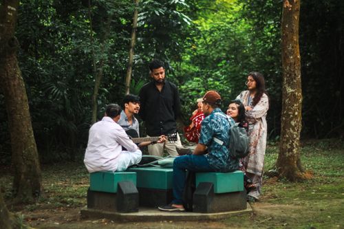 Free Friends gather in Dhaka park, enjoying music and nature in a lively outdoor setting. Stock Photo