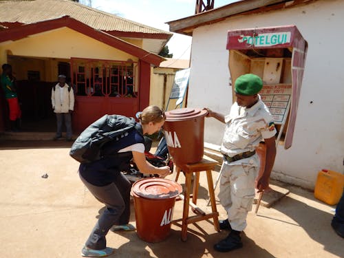 From above side view of unrecognizable female traveler with backpack washing hands from bucket on stool near African male in uniform standing on concrete pavement in summer