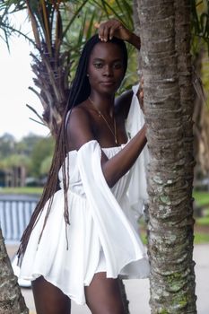 A woman in a white dress poses gracefully by trees in a natural setting, exuding elegance.