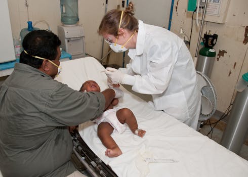 Doctor in protective gear treating a baby on a hospital bed with a parent present.