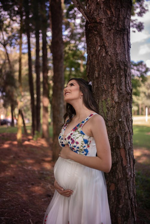 Free Woman in White and Blue Floral Spaghetti Strap Dress Standing Near Brown Tree Stock Photo