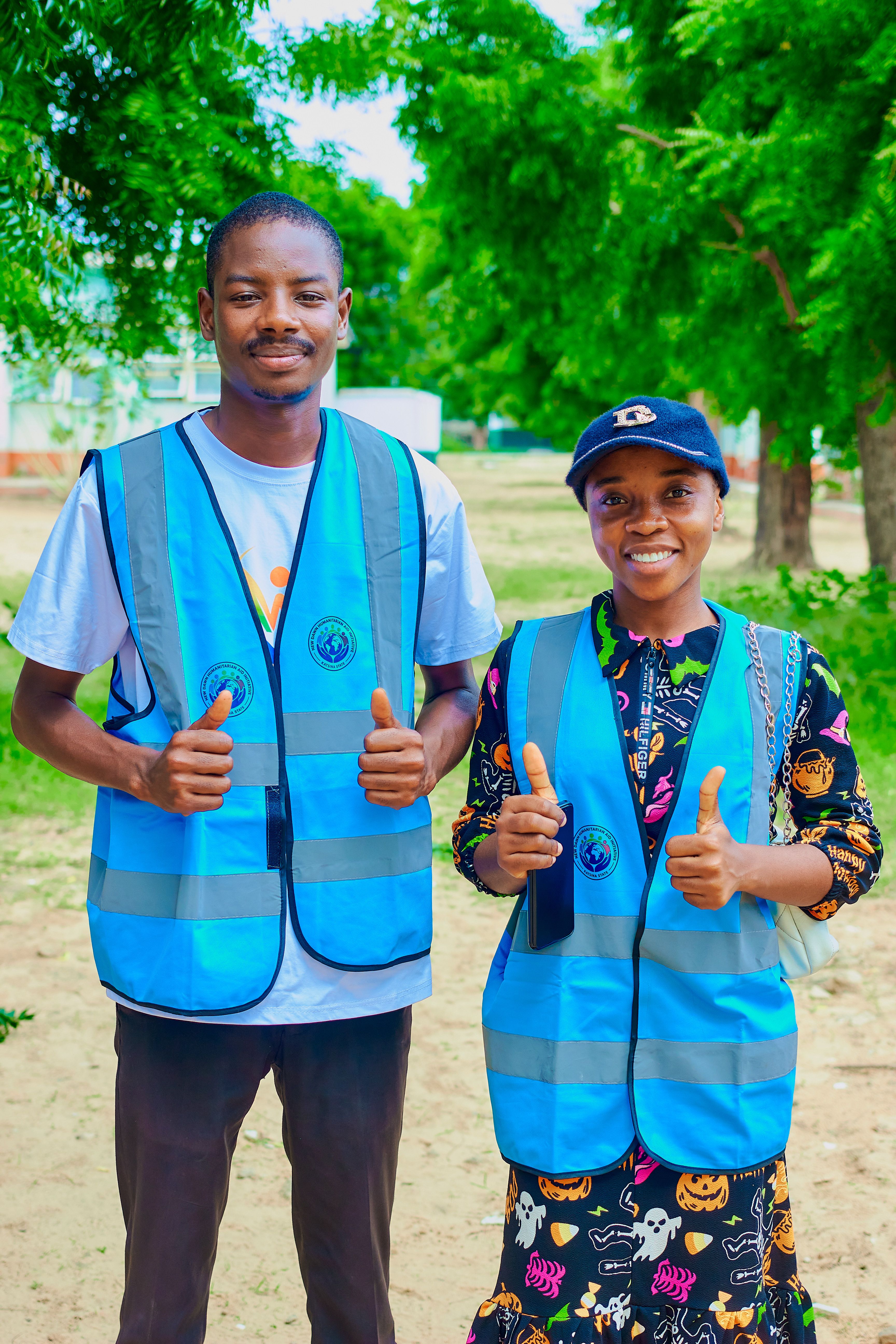 Two cheerful volunteers outdoors in Katsina, Nigeria, wearing blue vests, giving thumbs up.