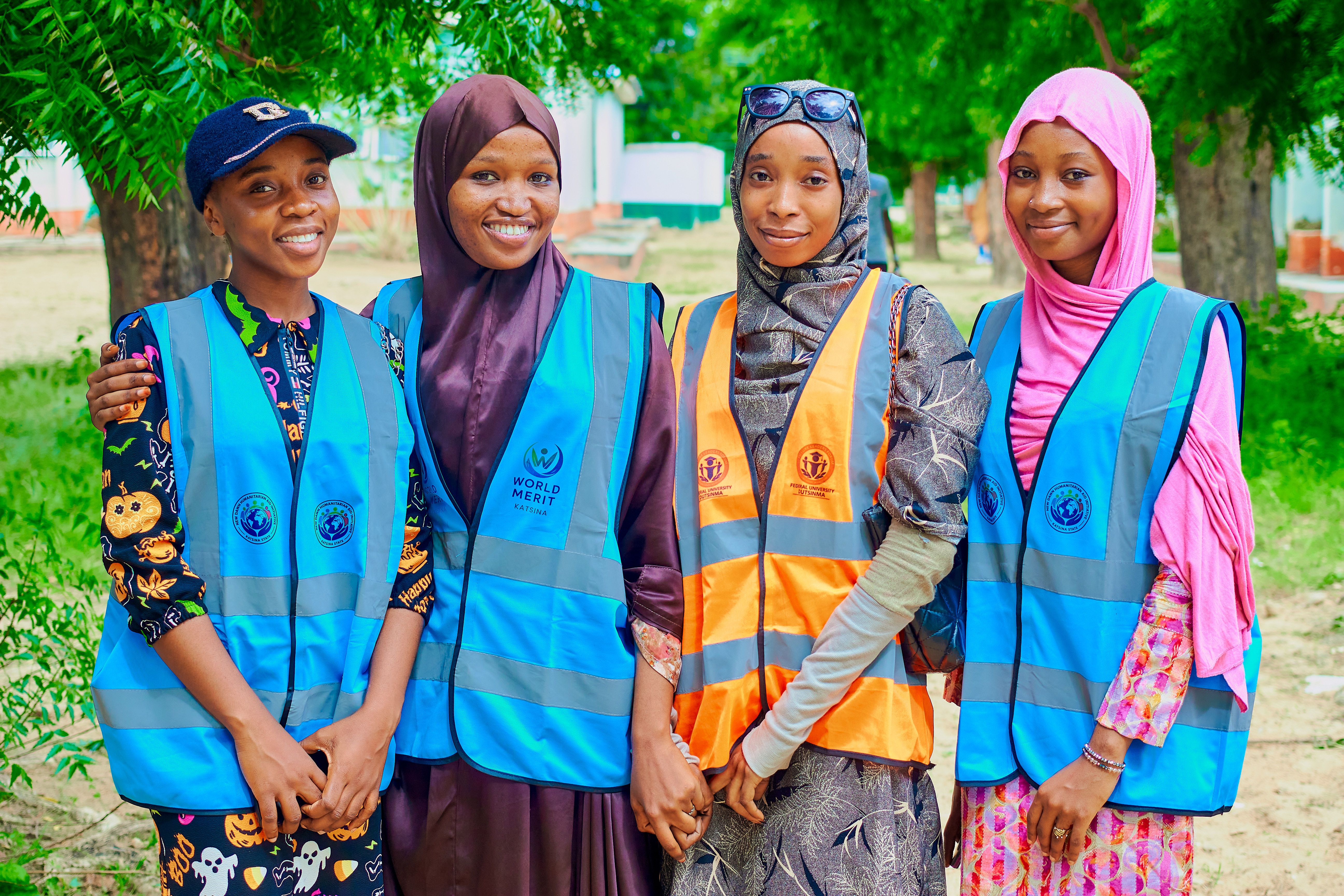 Four young volunteers in Katsina, Nigeria, wearing safety vests, smiling outdoors surrounded by trees.
